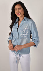 Load image into Gallery viewer, Front close up view of a woman wearing white pants and the frank lyman blue amore shirt. This shirt has a button down front, a drawstring hem with a tie in the middle, and two front patch pockets. The right pocket has sequins on it while the left pocket has words about love written on it.
