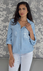 Load image into Gallery viewer, Front top half view of a woman wearing white pants and the frank lyman blue amore shirt. This shirt has a button down front, a drawstring hem with a tie in the middle, and two front patch pockets. The right pocket has sequins on it while the left pocket has words about love written on it.
