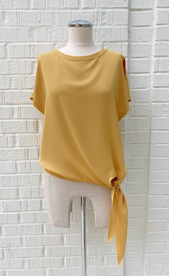 Front view of a mannequin wearing a marigold woven top from Frank Lyman with short sleeves and a front side tie at the bottom.