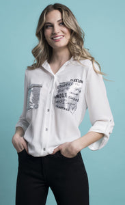 Front view of a woman with her hands in the pockets of her black pants and wearing the white amore top from Frank Lyman. This button down top features a front tie and two front sequin patch pockets. The left patch pocket has black lettering on it with words that say passion, amore, eternal, sweet, and more.