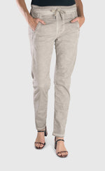Load image into Gallery viewer, Front bottom half view of a woman wearing the alembika grey iconic stretch jeans. This light grey jeans have a drawstring waistband with a tie, two front pockets and a relaxed slim/straight cut.
