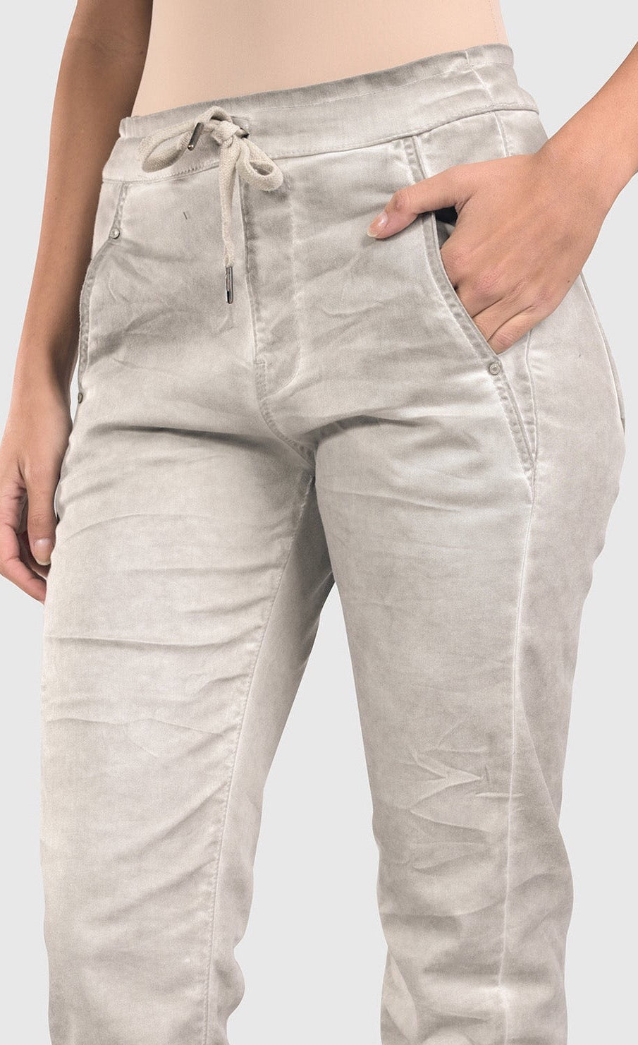 Front bottom half close up view of a woman wearing the alembika grey iconic stretch jeans. These light grey jeans have a drawstring waistband with a tie, two front pockets, and a relaxed slim/straight cut.