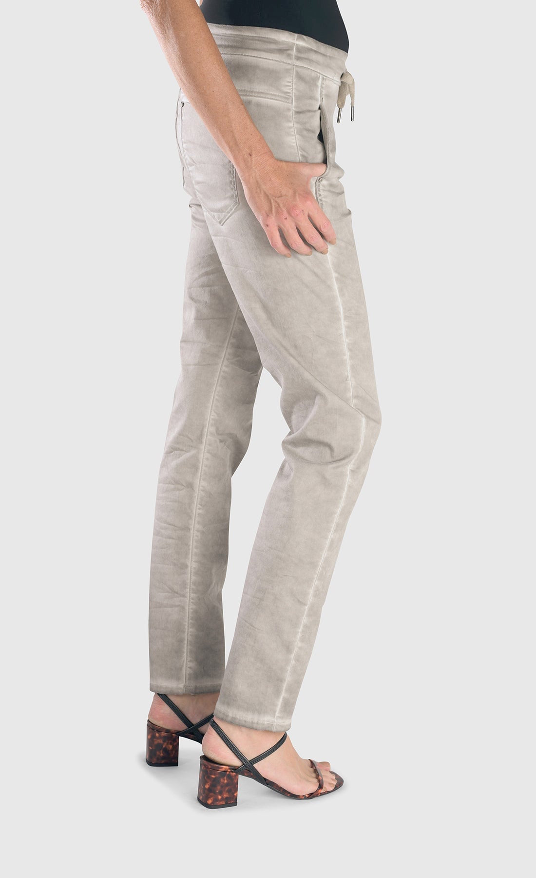 Right side bottom half view of a woman wearing the alembika grey iconic stretch jeans. These light grey jeans have a drawstring waistband with a tie, two front and back pockets and a relaxed slim/straight cut.
