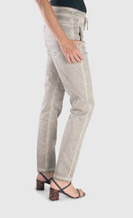 Load image into Gallery viewer, Right side bottom half view of a woman wearing the alembika grey iconic stretch jeans. These light grey jeans have a drawstring waistband with a tie, two front and back pockets and a relaxed slim/straight cut.
