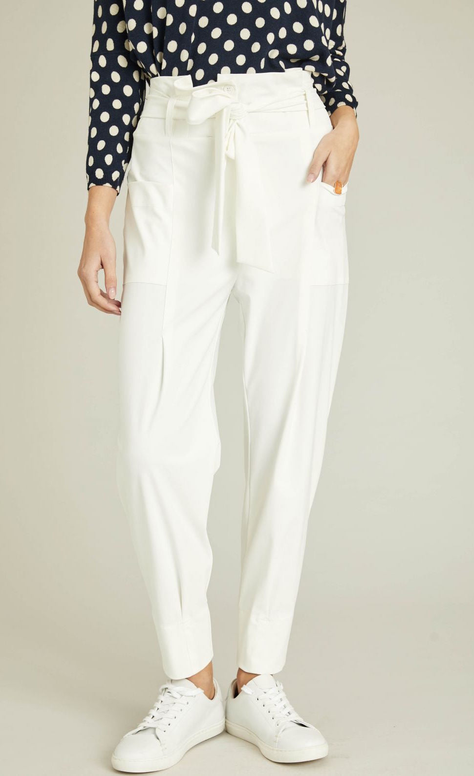 Front bottom half view of a woman wearing a blue and white polka dot top and the indies white collin pants. These pants have a carrot shape, two front patch pockets, and a tie belt.