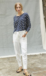 Load image into Gallery viewer, Front full body view of a woman wearing a blue and white polka dot top and the indies white collin pants. These pants have a carrot shape, two front patch pockets, and a tie belt.
