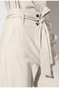 Front close up view of a woman wearing the Indies Collin Pant. This pant is pebble/beige colored. The wide waistband is high rise and has a tie belt. The side of the pants have pockets.