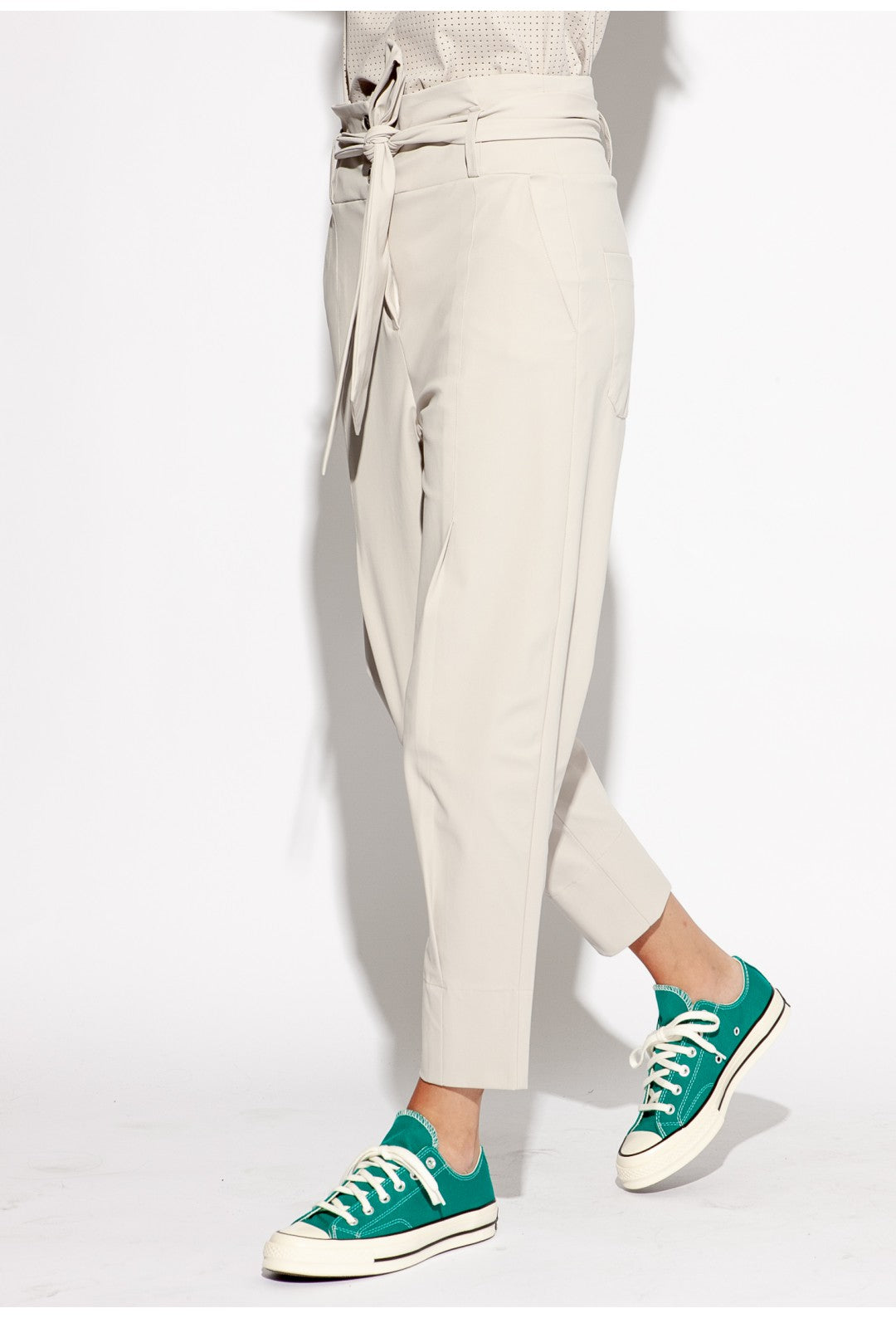 Front left sided bottom half view of a woman wearing the Indies Collin Pant. This pant is pebble/beige colored with a straight leg and a high rise waistband with a belt.