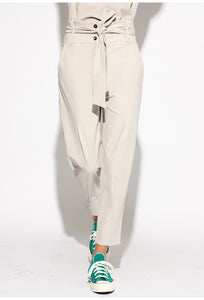 Front bottom half view of a woman wearing the Indies Collin Pant. This pant is pebble/beige colored with a straight leg and a high rise waistband with a belt.