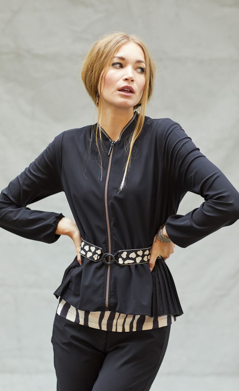 Front top half view of a woman wearing the indies black fute jacket. This jacket has a zip front, long sleeves, a short collar, and a belt with pink hearts around the waist.