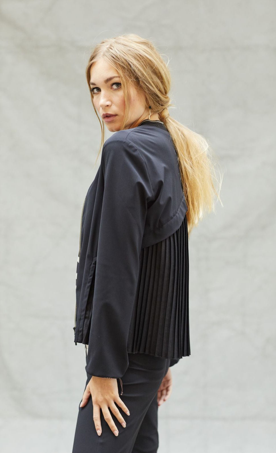 Back left side top half view of a woman wearing black pants and the indies black fute jacket. This jacket has pleats on the back with a criss-crossing of fabric on the upper back.