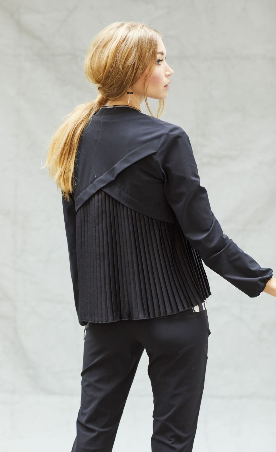 Back top half view of a woman wearing black pants and the indies black fute jacket. This jacket has pleats on the back with a criss-crossing of fabric on the upper back.