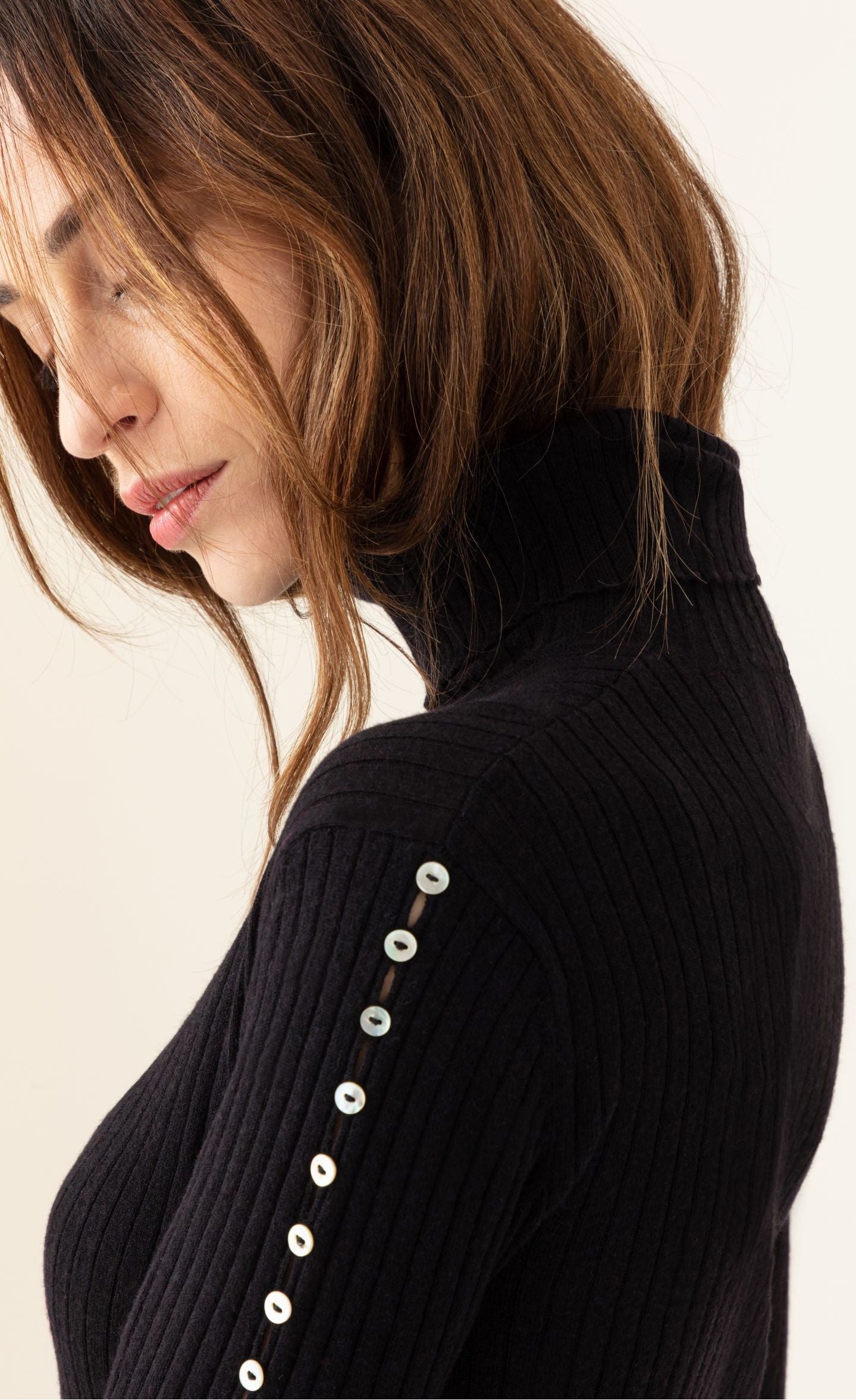Left side close up view of a woman wearing the fitted indies kris sweater. This ribbed turtleneck sweater is black with long sleeves that have pearl buttons running down the side.