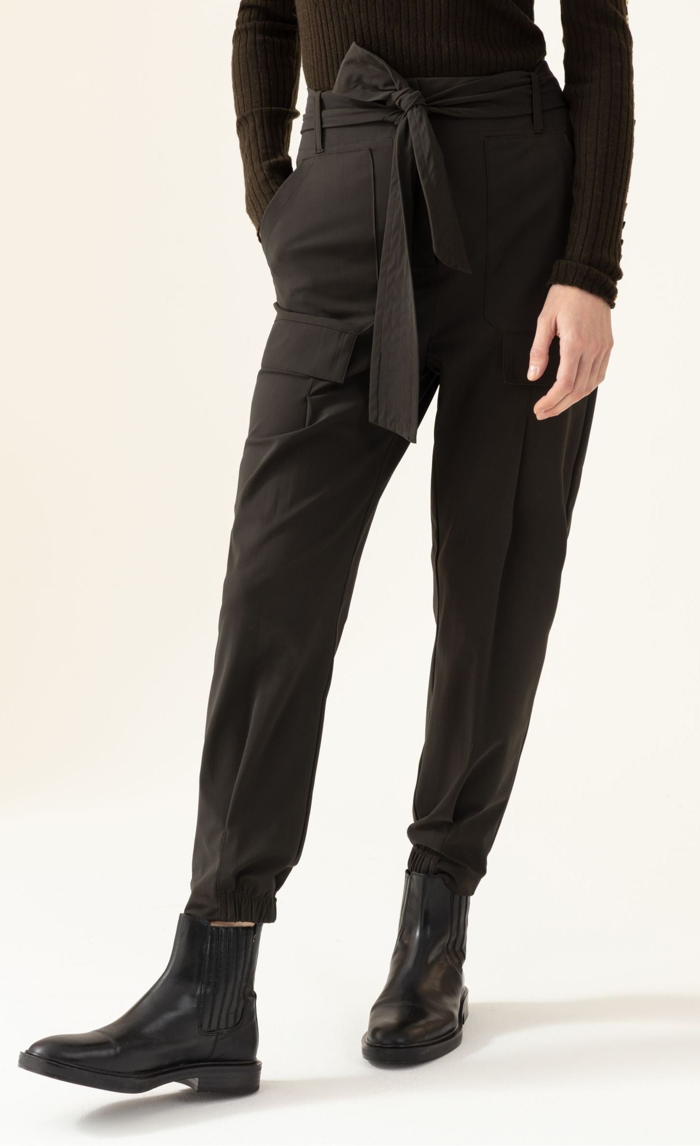Front bottom half view of a woman wearing the indies luna kaki pant. These pants have a large waistband with a tie belt, front patch pockets, a relaxed fit, and a tapered elastic hem.
