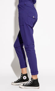 Left side bottom half view of a woman wearing the indies nico pant in the color indigo. This angle shows one of the two front patch pockets. The pant also has seams on the knees, back patch pockets, and a cuffed hem that ends above the ankles.