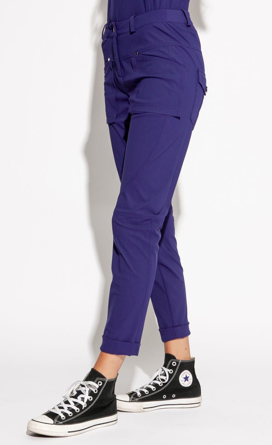 Front, left side bottom half view of a woman wearing the indies nico pant in the color indigo. This pant has two front patch pockets. The pant also has seams on the knees, back patch pockets, and a cuffed hem that ends above the ankles.