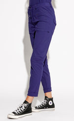 Load image into Gallery viewer, Front, left side bottom half view of a woman wearing the indies nico pant in the color indigo. This pant has two front patch pockets. The pant also has seams on the knees, back patch pockets, and a cuffed hem that ends above the ankles.
