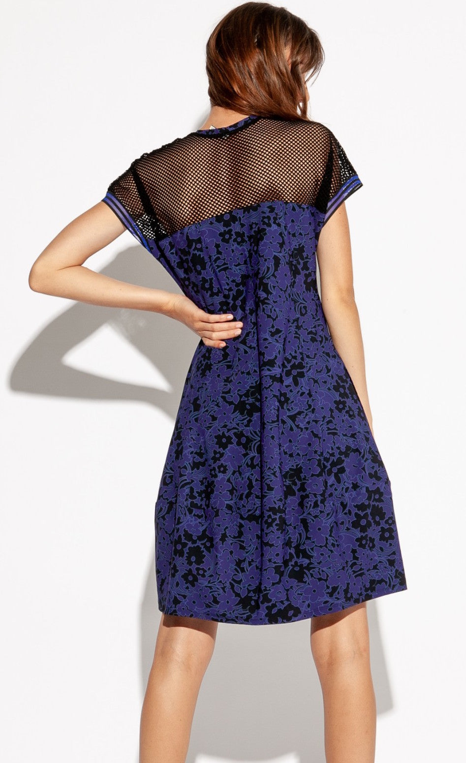Back full body view of a woman with her left hand on her hip and wearing the Indies Skipy Dress. This dress is indigo blue and black with a floral print and black mesh on the shoulders and short sleeves. The dress is loose fitting with a slightly fitted waist and a cut that sits above the knees.