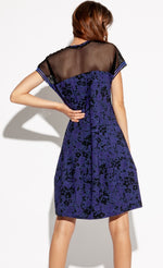 Load image into Gallery viewer, Back full body view of a woman with her left hand on her hip and wearing the Indies Skipy Dress. This dress is indigo blue and black with a floral print and black mesh on the shoulders and short sleeves. The dress is loose fitting with a slightly fitted waist and a cut that sits above the knees.
