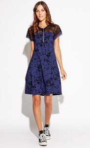 Front full body view of a woman wearing the Indies Skipy Dress. This dress is indigo blue and black with a floral print and black mesh on the shoulders and short sleeves. The dress is loose fitting with a slightly fitted waist and a cut that sits above the knees.