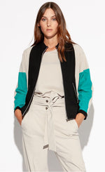 Load image into Gallery viewer, Front top half view of a woman with her hands in the pockets of the pebble colored indies Collin Pant. On the top she is wearing the pebble colored, perforated Indies cara tee shirt underneath the Indies Nano Jacket. The jacket has black, seafoam blue, and pebble/beige color blocking. The sleeves are seafoam green, the front zip down front is surrounded by black, and the shoulders and side are pebble/beige. The jacket the has two front zipper pockets.
