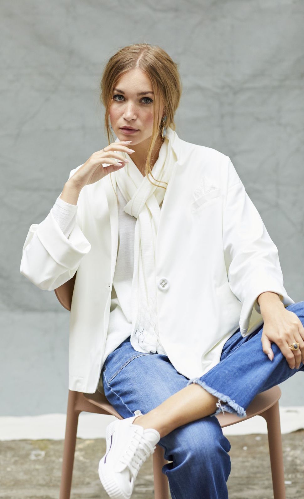 Front view of a woman sitting on a chair wearing blue jeans and the white indies voyageur jacket. This jacket is open and layered over a white top with a with scarf. The jacket has a single front button.