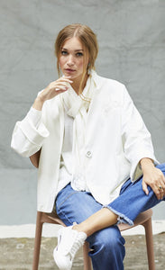 Front view of a woman sitting on a chair wearing blue jeans and the white indies voyageur jacket. This jacket is open and layered over a white top with a with scarf. The jacket has a single front button.