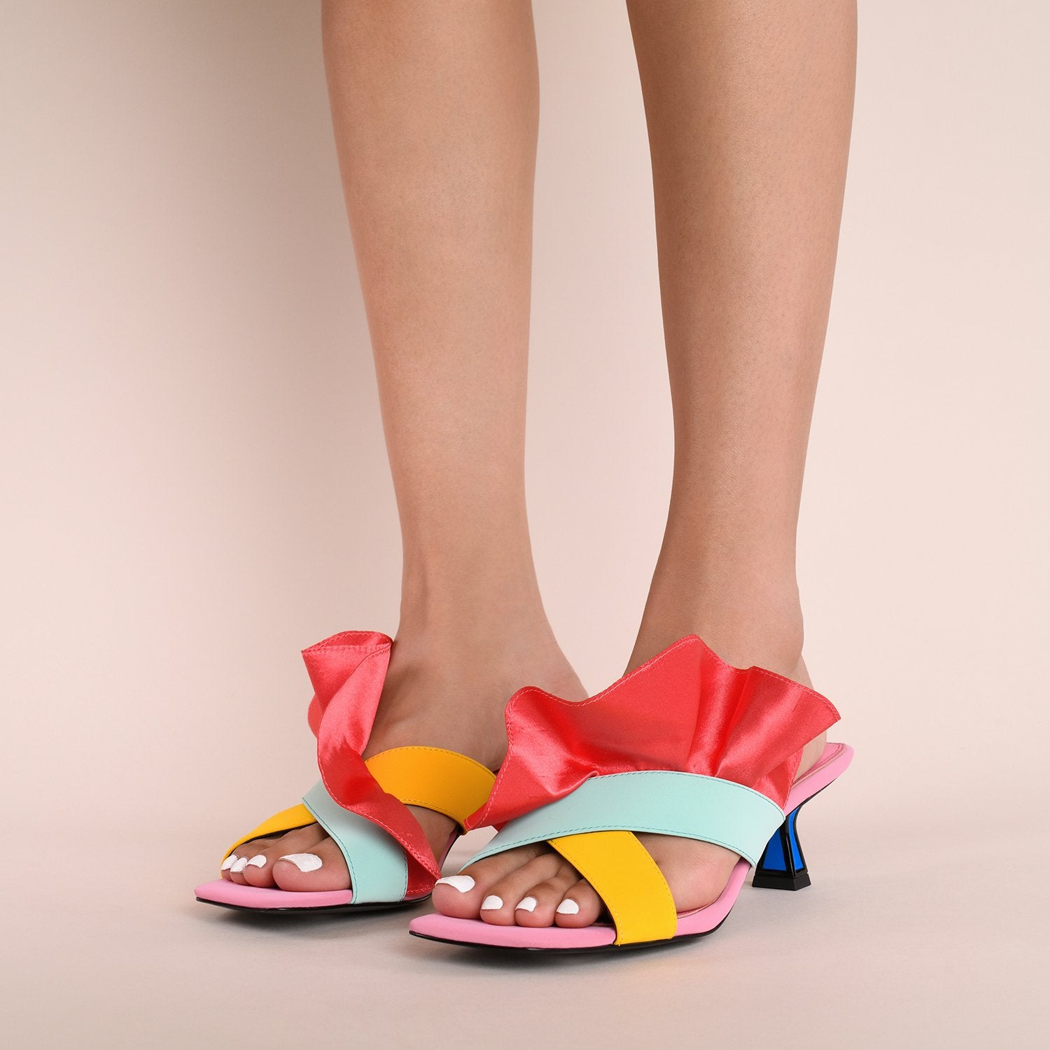 Front outer side view of a model wearing a pair of the kat maconie dia kitten heel. This slip on heel features a pink sole, a tiny blue heel outlined with black, two cross over straps, and a pink/red satin ruffle attached to the upper.