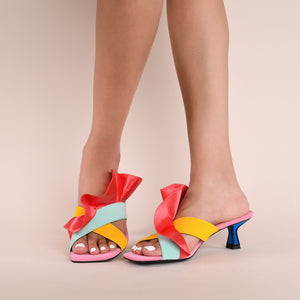 Front and inner side view of a model wearing a pair of the kat maconie dia kitten heel. This slip on heel features a pink sole, a tiny blue heel outlined with black, two cross over straps, and a pink/red satin ruffle attached to the upper.