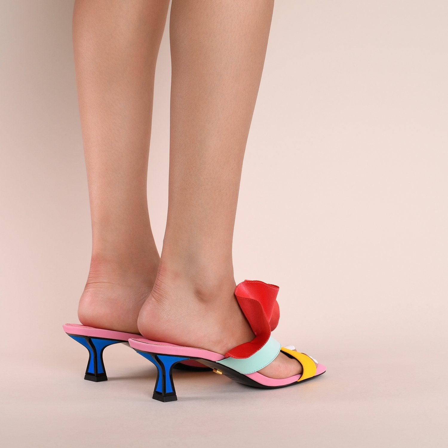 Back outer side view of a model wearing a pair of the kat maconie dia kitten heel. This slip on heel features a pink sole, a tiny blue heel outlined with black, two cross over straps, and a pink/red satin ruffle attached to the upper.