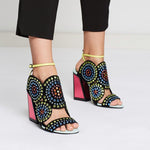 Load image into Gallery viewer, Outer and inner side view of a model wearing a pair of the kat maconie frida multibrights high heels. This shoe is black with multicolored rhinestones arranged in circles. The toe and back are open. The shoe features a yellow ankle strap and a pink high heel with a black outline.
