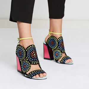 Outer and inner side view of a model wearing a pair of the kat maconie frida multibrights high heels. This shoe is black with multicolored rhinestones arranged in circles. The toe and back are open. The shoe features a yellow ankle strap and a pink high heel with a black outline.