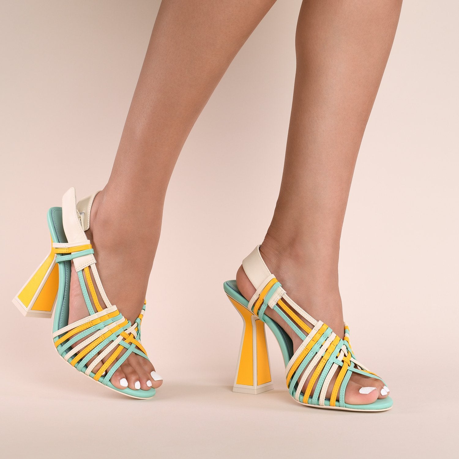 Front inner and outer side views of a model wearing a pair of the kat maconie sheri sandal. This strappy sandal has a high heel and a mix of turquoise, yellow, and white colored straps. The white back strap is wider and adjustable. The heel is yellow and outlined with opal/white.