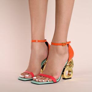 Outer side view of a model wearing a pair of the kat maconie suzu high heel. This shoe has a blue/opal sole, an orange and pink patent leather upper with an adjustable ankle strap, and a gold chain-like heel.