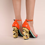 Load image into Gallery viewer, Back view of a model wearing a pair of the kat maconie suzu high heel. This shoe has a blue/opal sole, an orange and pink patent leather upper with an adjustable ankle strap, and a gold chain-like heel.

