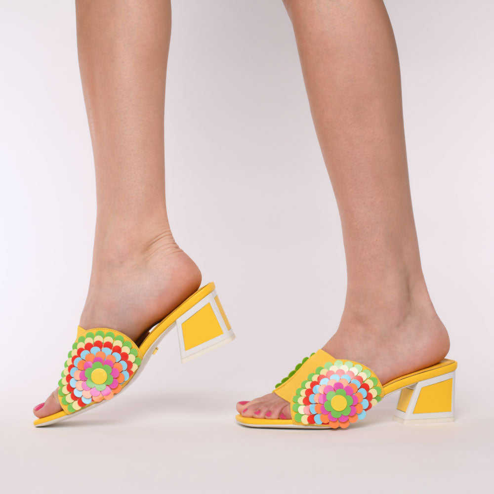 side view of a woman wearing a pair of the kat maconie vira kicker heel sandals. These mule sandals are mineral yellow with multi colored flowers on the sides. These sandals also have a block heel and an open toe front.