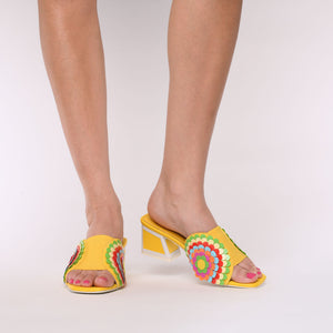 Front view of a woman wearing a pair of the kat maconie vira kicker heel sandals. These mule sandals are mineral yellow with multi colored flowers on the sides. These sandals also have a block heel and an open toe front.
