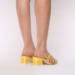 Load image into Gallery viewer, Back view of a woman wearing a pair of the kat maconie vira kicker heel sandals. These mule sandals are mineral yellow with multi colored flowers on the sides. These sandals also have a block heel and an open toe front.
