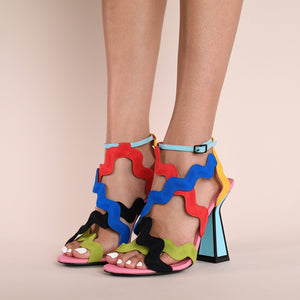 Front, outer side view of a model wearing a pair of the kat maconie jihan high heel. This sandal shoe is made up of multi-colored, wave-like embroidered upper straps. The shoe features a blue ankle strap and a matching blue heel outlined with black.