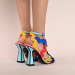 Load image into Gallery viewer, Back, outer side view of a model wearing a pair of the kat maconie jihan high heel. This sandal shoe is made up of multi-colored, wave-like embroidered upper straps. The shoe features a blue ankle strap and a matching blue heel outlined with black.
