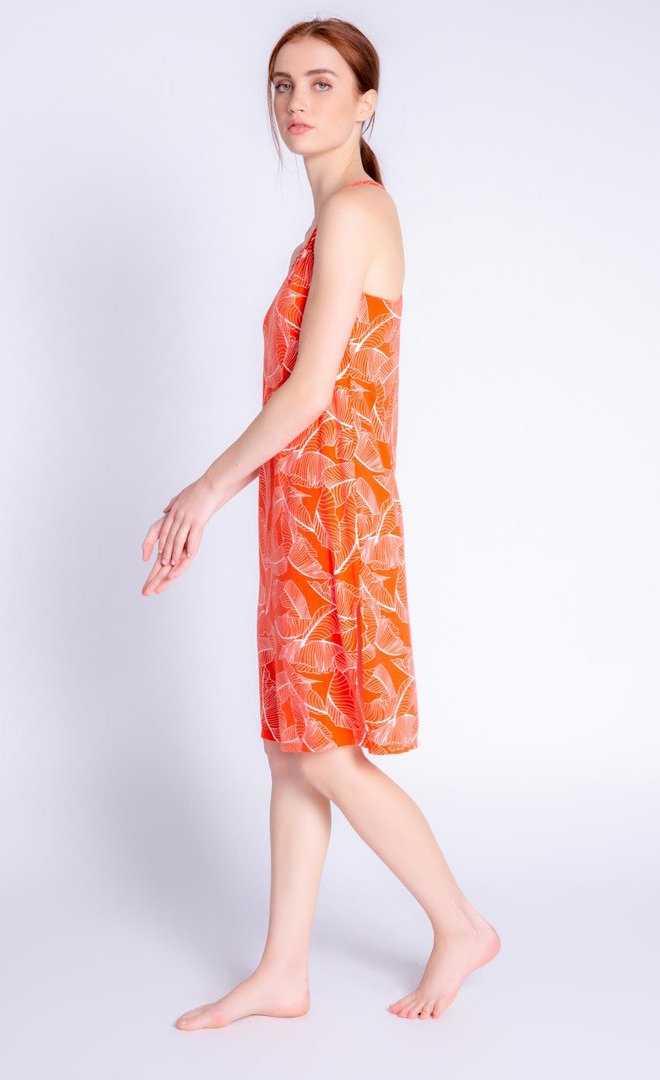 Left side full body view of a woman wearing the pj salvage leafy dreams dress. This tank dress has a v-neck, long side slits, and is orange colored with a white tropical leaf print.