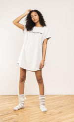 Load image into Gallery viewer, Front view of a woman wearing the coffee shoppe t-shirt dress. This t-shirt dress is grey with the words let&#39;s sleep in.
