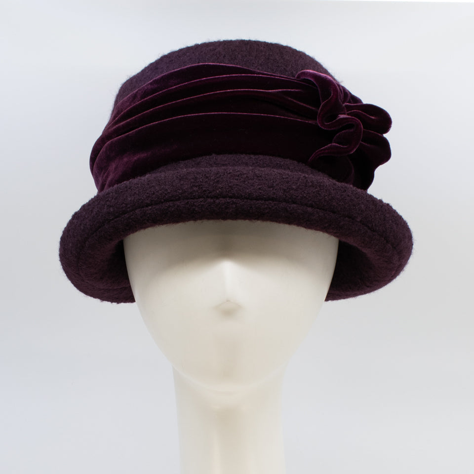 Front view of the lillie & cohoe aubergine wool jeanette hat. This hat is wine/purple colored and has a rounded brim and a velvet band that wraps around the crown to make a bow on the side.