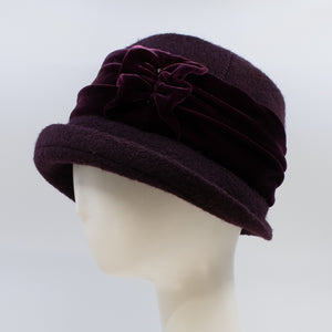 left side view of the lillie & cohoe aubergine wool jeanette hat. This hat is wine/purple colored and has a rounded brim and a velvet band that wraps around the crown to make a bow on the side.