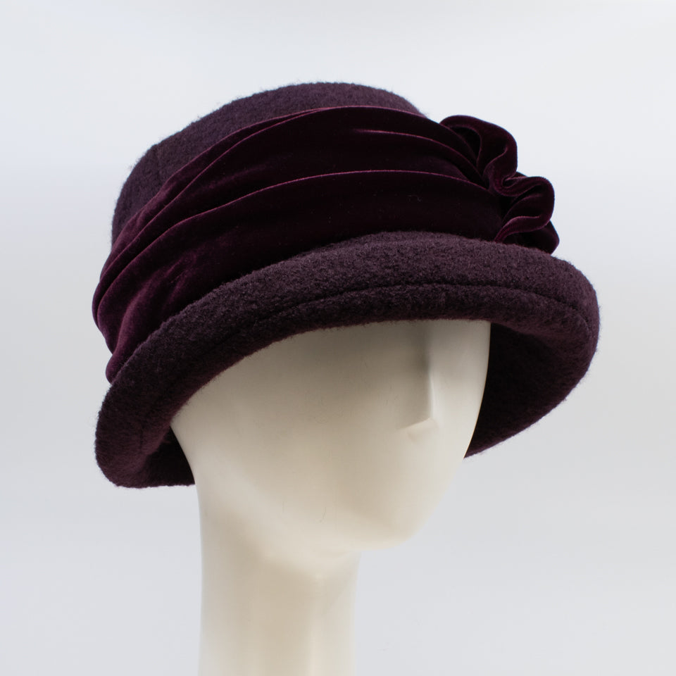 Front right side view of the lillie & cohoe aubergine wool jeanette hat. This hat is wine/purple colored and has a rounded brim and a velvet band that wraps around the crown to make a bow on the side.