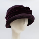 Load image into Gallery viewer, Front right side view of the lillie &amp; cohoe aubergine wool jeanette hat. This hat is wine/purple colored and has a rounded brim and a velvet band that wraps around the crown to make a bow on the side.
