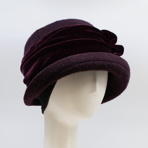 left side view of the lillie & cohoe aubergine wool jeanette hat. This hat is wine/purple colored and has a rounded brim, ear warmers, and a velvet band that wraps around the crown to make a bow on the side.