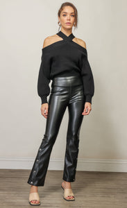 Front full body view of a woman wearing the Line & Dot Cold Shoulder sweater tucked into black leather pants. This black sweater has a criss cross neck that exposes the shoulder and long sleeves.