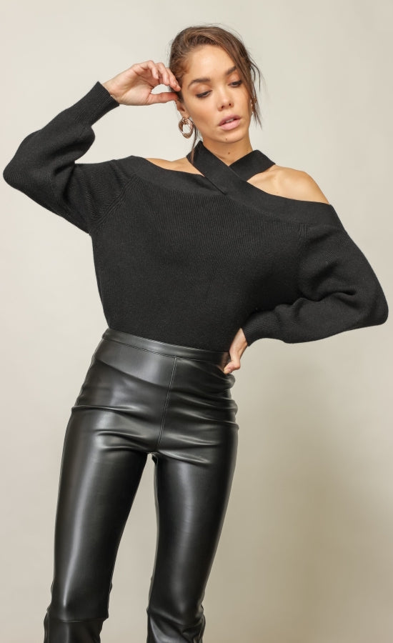 Front top half view of a woman with one hand on her hip and one hand by her forehead wearing the Line & Dot Cold Shoulder sweater tucked into black leather pants. This black sweater has a criss cross neck that exposes the shoulder and long sleeves.