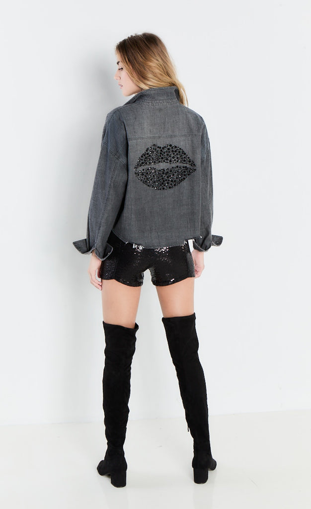 back full body view of a woman wearing the lisa todd black denim back talk shirt. This back of this shirt is decorated with stones that take the shape of lips. The shirt has long sleeves and a distressed hem.